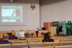 Aula z rzędami krzeseł, na których siedzą osoby. Na środku katedra z mównicą. Z jej prawej strony stoi kobieta z mikrofonem. Za katedrą ekran projekcyjny.