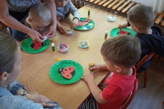 Siedząca grupa  dzieci przy drewnianym stoliku i wykonująca pracę plastyczną - biedronki na zielonych talerzykach.