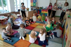 Siedzące na dywanie przedszkolaki i Pani, które z nimi rozmawia, a w tle cztrey stojące Panie, które obserwują spotkanie.