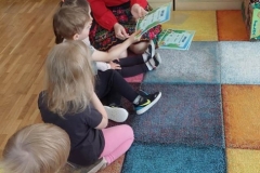 Siedząca grupa dzieci na dywanie, patrząca na Panią w czerwonej bluzce, która rozdaje dyplomy wzorowego czytelnika.