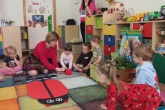 Siedząca grupa dzieci na dywanie, patrząca na Panią w czerwonej bluzce, która rzuca kostką.