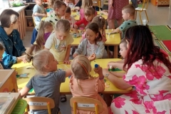 Siedząca grupa dzieci przy żółtym stoliku i wykonująca prace plastyczne.