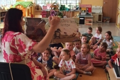 Sala przedszkolna, osoba trzymająca książkę w rękach i wpatrzone w nią dzieci, które siedzą na podłodze.