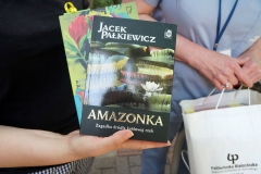 Po lewej osoba trzymająca w dłoni dwie książki, po prawej widoczne ręce kobiety trzymającej torebkę upominkową.