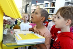 Chłopiec wpatrzony w ekran urządzenia do zwrotu i wypożyczeń książek. W tle grupa dzieci w wieku przedszkolnym.