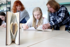 Na pierwszym planie książka wykonana w technice book origami prezentująca kształt serca. W tle osoby biorące udział w warsztatach.