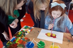 Dziewczynka wykonuje pracę plastyczną. Nad nią pochylają się dwie kobiety.