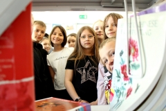 Dziewczynki i chłopcy spoglądają w obiektyw aparatu fotograficznego zza półek regału z książkami