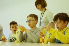 Trzej chłopcy naklejają żółtą bibułę na rolkę papieru. Za nimi stoi kobieta w białej koszulce