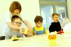 Przy stole siedzą trzej chłopcy. Jednemu z nich przy naklejaniu pomaga kobieta. Na blacie leżą nożyce, klej, kawałki kolorowej bibuły