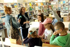 Grupa dzieci patrzy się na kobietę, która coś mówi. W tle ściany z regałami wypełnionymi kolorowymi publikacjami