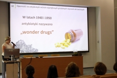 Kobieta z mikrofonem stoi z lewej strony katedry, nad którą znajduje się duży ekran z prezentacją o tematyce związanej z antybiotykami.