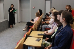 Z lewej strony kobieta, dyrektor Biblioteki PB, trzymająca w ręku mikrofon i zieloną teczkę,   przemawia do siedzących na auli osób.