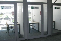 Widok ławek i dosuniętych do nich krzeseł w przeszklonych pokojach pracy.