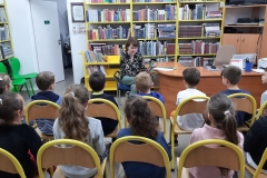 Młoda dziewczyna siedzi naprzeciwko grupki dzieci usadowionych na krzesłach i czyta im książkę.