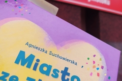 Książka Agnieszki Suchowierskiej "Miasto ze złotym sercem". W tle czerwona tablica o z nazwą szkoły.
