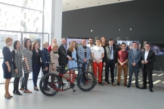 Grupa organizatorów i ambasadorów kampanii "Sam zadbaj o swoje bezpieczeństwo" stoi w szeregu. Przed nimi znajduje się czarno-czerwony rower. 