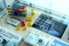 Gablota, a w niej znajduje się aparat z klocków lego, kilka pojedynczych klocków lego i mini figurka, aparaty cyfrowe, zdjęcie kolorowe, książki.
