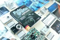 Pięć różnorodnych aparatów cyfrowych. Obok aparatów znajdują się kartki z informacjami o aparatach, książki i zdjęcia.