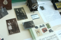 Widok z góry na fragment gabloty. Widoczny aparat Agfa Prontor II, stare czarno-białe zdjęcia, szklane negatywy, książka i zapisane kartki papieru.