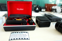 Na pierwszym planie widać aparat fotograficzny Rollei A110 Tessar, z prawej strony czarne futerały, w tle inne aparaty fotograficzne.