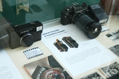 Fragment gabloty z dwoma aparatami fotograficznymi Praktika i Elikon. Obok stare zdjęcia czarno-białe i kartka z  informacją o aparacie w kształcie książki.
