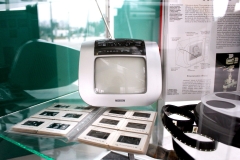 Szklana gablota z małym telewizorem dawnego typu. Z przodu album z kliszą na rzutnik i stary film na szpuli. W tle otwarta książka z opisem działania telewizora.