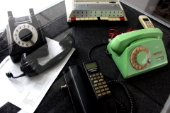  Trzy dawne telefony. Zielony i czarny z tarczą do wykręcania numeru oraz czarny telefon samochodowy.