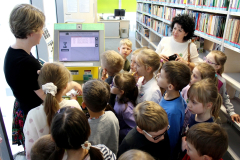 Dzieci zgromadzone przy stanowisku w bibliotece obserwują działanie urządzenia związanego z obsługą książek