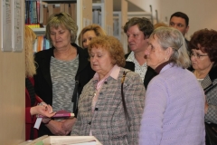 Grupa starszych ludzi słucha oprowadzającej kobiety w czerwonej marynarce, która trzyma w dłoni książki i pokazuje zebranym.