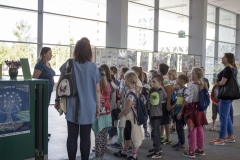 W przeszklonym holu stoi tyłem kilkunastoosobowa grupa dzieci. Przed dziećmi stoi kobieta, które opowiada coś dzieciom.