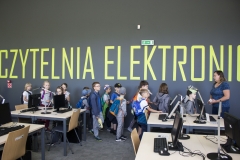 Za rzędami stanowisk komputerowych duża grupa dzieci. Przed nimi kobieta w niebieskiej bluzce i jeansach. Na ciemnoszarej ścianie żółty napis - czytelnia elektroniczna. Nad napisem zielona tabliczka z kierunkiem ewakuacji.