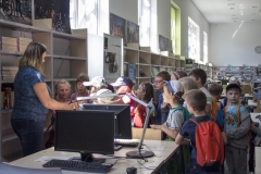 Czternastoosobowa grupa dzieci otacza kobietę stojącą przy biurku, która pokazuje dzieciom książkę. Za dziećmi widok na pomieszczenie czytelni.