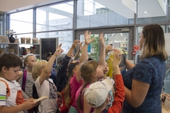 Dziesięcioosobowa grupa dzieci stoi w pomieszczeniach czytelni książek i podnosi ręce do góry. Przed nimi stoi kobieta z książką w ręku.